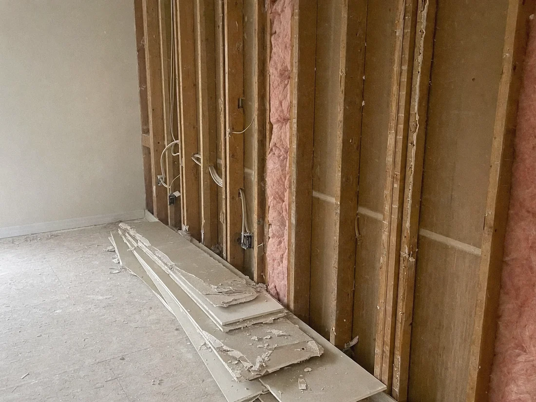 Drywall stripped from a wall exposing studs, wiring and insulation, with old board stacked on the floor